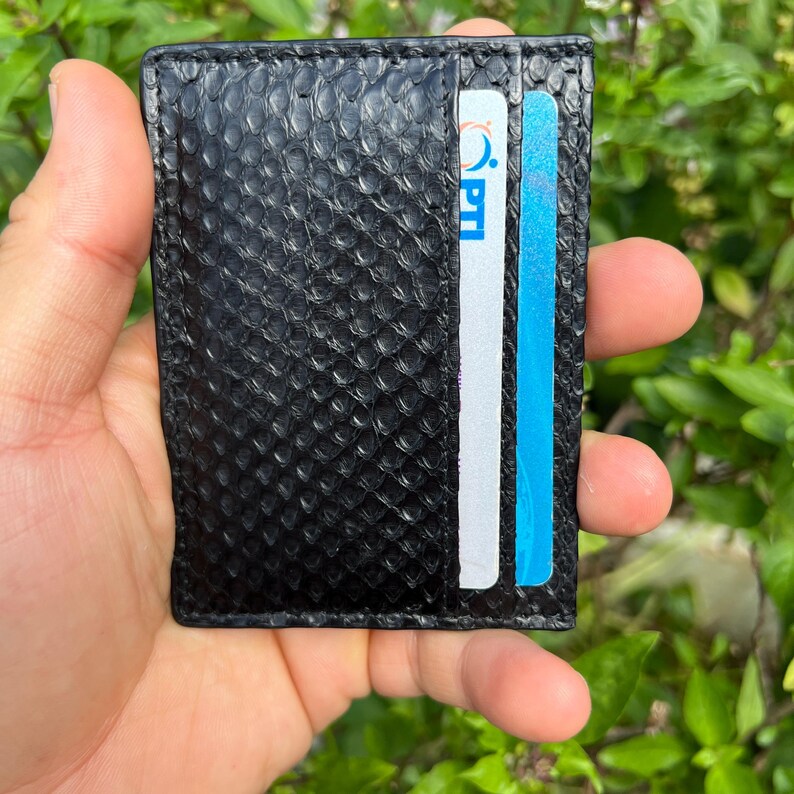 Puede incluir: Tarjetero de cuero negro con un patr&oacute;n de piel de serpiente texturizado. La cartera tiene dos ranuras para tarjetas, una con una tarjeta blanca y otra con una tarjeta azul. El tarjetero se sostiene en una mano.