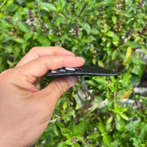 May include: A black, textured, rectangular object with small, colorful squares on the top surface. The object is held in a hand against a backdrop of green foliage. The object appears to be a small wallet or card holder.
