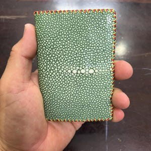 Könnte beinhalten: Eine grüne Brieftasche aus Stachelrochenleder mit einem einzigartigen strukturierten Muster. Die Kanten sind mit rotem, gelbem und goldenem Faden genäht. Die Brieftasche wird in der Hand einer Person gehalten.