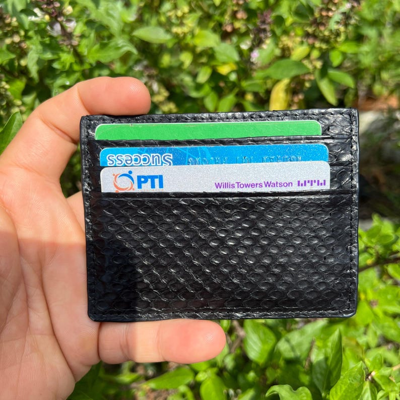 Puede incluir: Un tarjetero negro con un patr&oacute;n texturizado similar a la piel de serpiente. El tarjetero contiene varias tarjetas con texto visible, incluyendo "SUCCESS", "PTI" y "Willis Towers Watson". El fondo es follaje verde borroso.