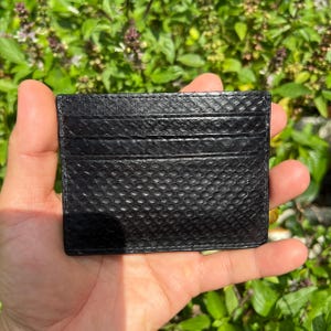 Puede incluir: Un tarjetero negro con un patr&oacute;n texturizado similar a la piel de serpiente. El tarjetero tiene m&uacute;ltiples ranuras para tarjetas y se sostiene en la mano de una persona contra un fondo de follaje verde.
