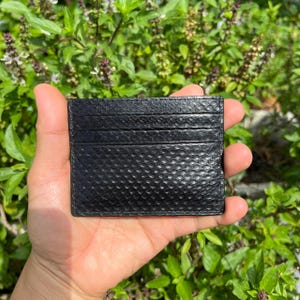 Puede incluir: Un tarjetero negro con un patr&oacute;n texturizado similar a la piel de serpiente. La cartera rectangular tiene m&uacute;ltiples ranuras para tarjetas y se sostiene en una mano sobre un fondo de follaje verde. La cartera es de color oscuro.
