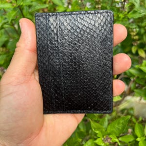 Puede incluir: Un tarjetero negro con un patr&oacute;n texturizado de piel de serpiente. El tarjetero tiene m&uacute;ltiples ranuras para tarjetas y est&aacute; sostenido en la mano de una persona. El fondo es follaje verde borroso.