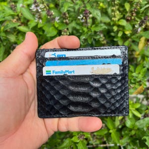 May include: Black leather card holder with a textured snakeskin pattern. The wallet is filled with several cards, including one with the FamilyMart logo. The background is a lush green with leafy foliage.