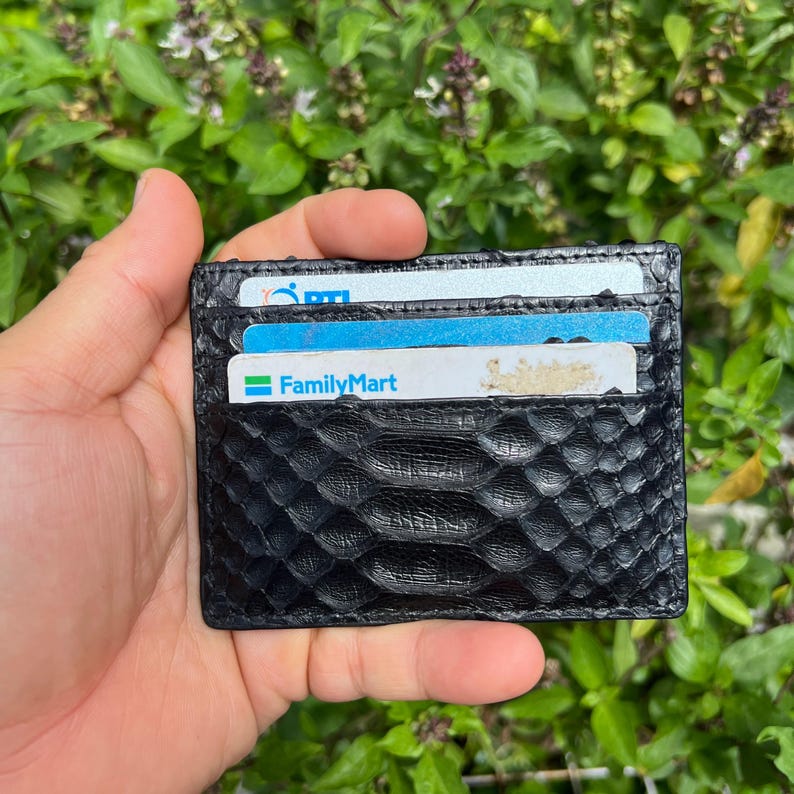 May include: Black leather card holder with a textured snakeskin pattern. The wallet is filled with several cards, including one with the FamilyMart logo. The background is a lush green with leafy foliage.