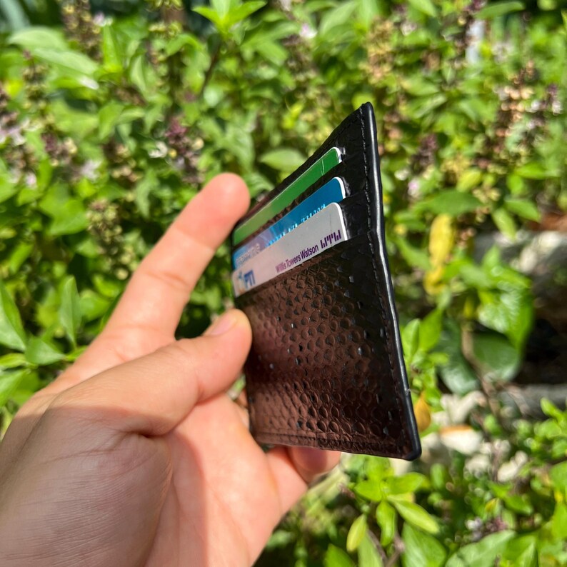 Puede incluir: Tarjetero de cuero negro con un patr&oacute;n texturizado, que contiene varias tarjetas con texto visible. El tarjetero se sostiene contra un fondo de follaje verde. Dise&ntilde;ado para guardar tarjetas de cr&eacute;dito y otras tarjetas esenciales.