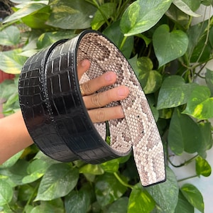 Pode incluir: Um cinto preto com textura de crocodilo no exterior e um padrão de pele de cobra no interior. O cinto é exibido em um fundo de folhas verdes.