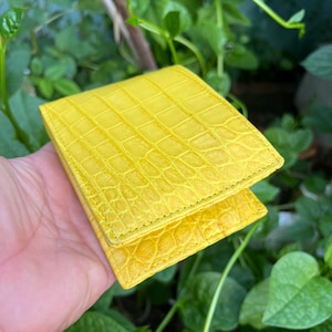 Può includere: Un portafoglio in pelle giallo brillante, di forma quadrata e con una superficie strutturata. Il portafoglio è tenuto in mano su uno sfondo di fogliame verde. La pelle presenta un motivo simile a quello di un rettile.