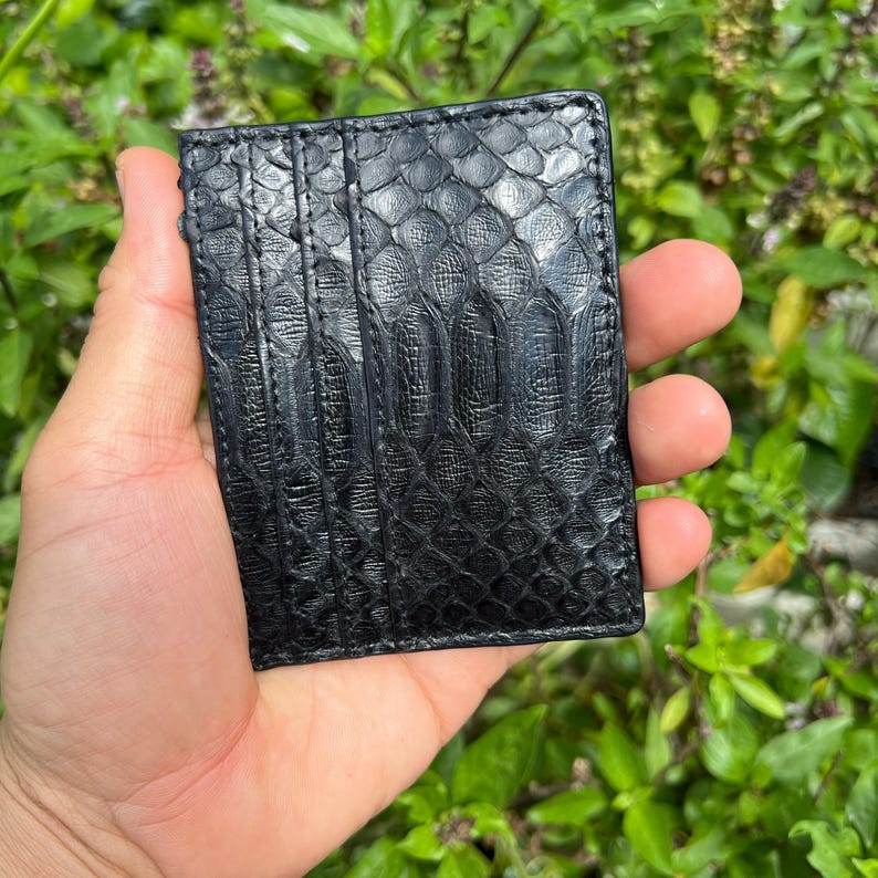 May include: Black card holder with a textured, snake-skin pattern. The wallet has multiple card slots and is held in a hand against a background of green foliage.