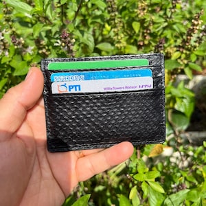 Könnte beinhalten: Schwarzer Lederkartenhalter mit strukturiertem Muster, der mehrere Karten enthält. Die Karten sind grün, blau und weiß, mit Text wie "SUCCESS", "PTI" und "Willis Towers Watson". Die Geldbörse wird vor grünem Laub gehalten.