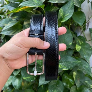 Könnte beinhalten: Ein schwarzer Gürtel mit einem strukturierten Schlangenhautmuster. Der Gürtel hat eine silberfarbene rechteckige Schnalle. Der Gürtel wird vor einem Hintergrund aus grünen Blättern gehalten.