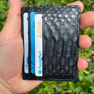 May include: Black card holder with a textured, snake-skin pattern. The wallet is open, revealing several credit cards. One card has the FamilyMart logo. The wallet is held in a person's hand against a background of green foliage.