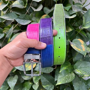 Puede incluir: Una colección de cinturones de cuero coloridos en rosa, morado, azul y verde. Los cinturones tienen un patrón texturizado, similar al de un reptil, y hebillas plateadas. Los cinturones se sostienen contra un fondo de follaje verde.