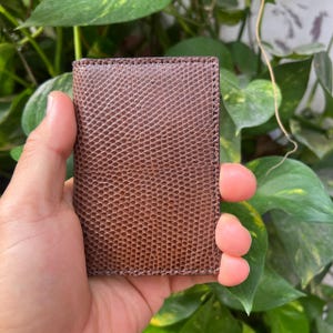 Può includere: Un porta carte rettangolare marrone caratterizzato da una texture goffrata che imita la pelle di lucertola. L'accessorio presenta bordi cuciti netti lungo il perimetro e un orientamento verticale sottile, con una finitura marrone scuro uniforme.