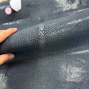 以下が含まれることがあります： 中央に自然な白い円形の模様がある、独特で凹凸のある粒状の質感を持つダークグレーのエイ革。素材にはわずかな光沢があり、数字の9が記された小さな円形のタグが付いています。