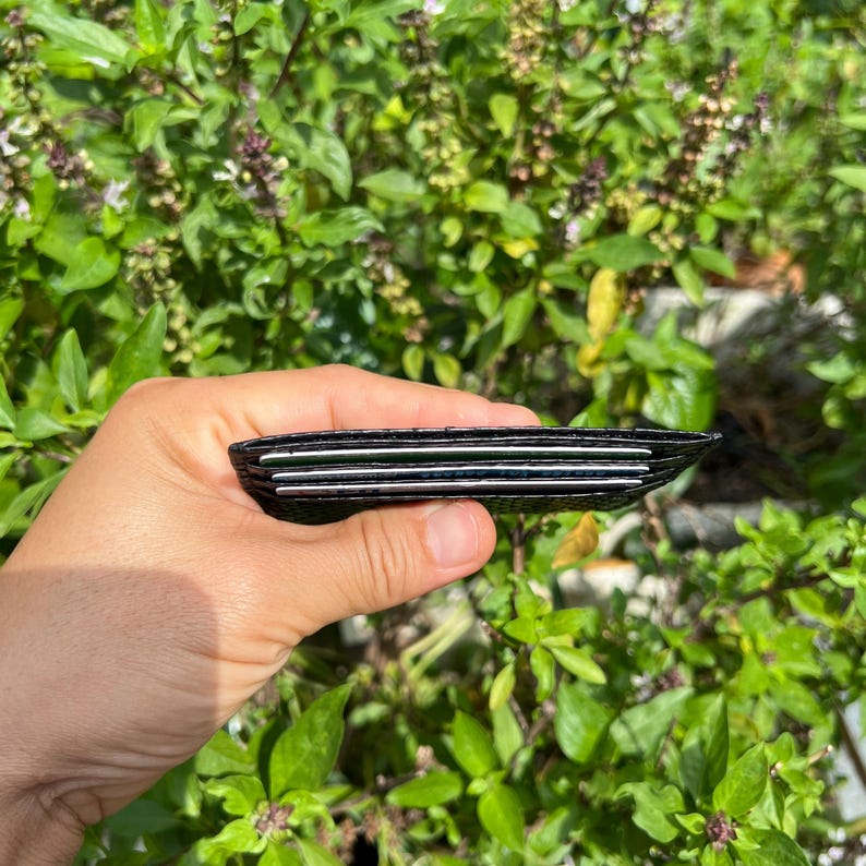 Puede incluir: Una cartera negra con m&uacute;ltiples ranuras para tarjetas se sostiene contra un fondo de follaje verde. La cartera parece estar hecha de material texturizado y contiene varias tarjetas visibles. La imagen est&aacute; tomada con luz natural.