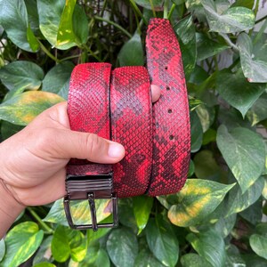 Puede incluir: Un cinturón rojo y negro con estampado de piel de serpiente y una hebilla plateada. El cinturón está hecho de un material texturizado y tiene varios agujeros para ajustar. La hebilla es rectangular con una sola hebilla.