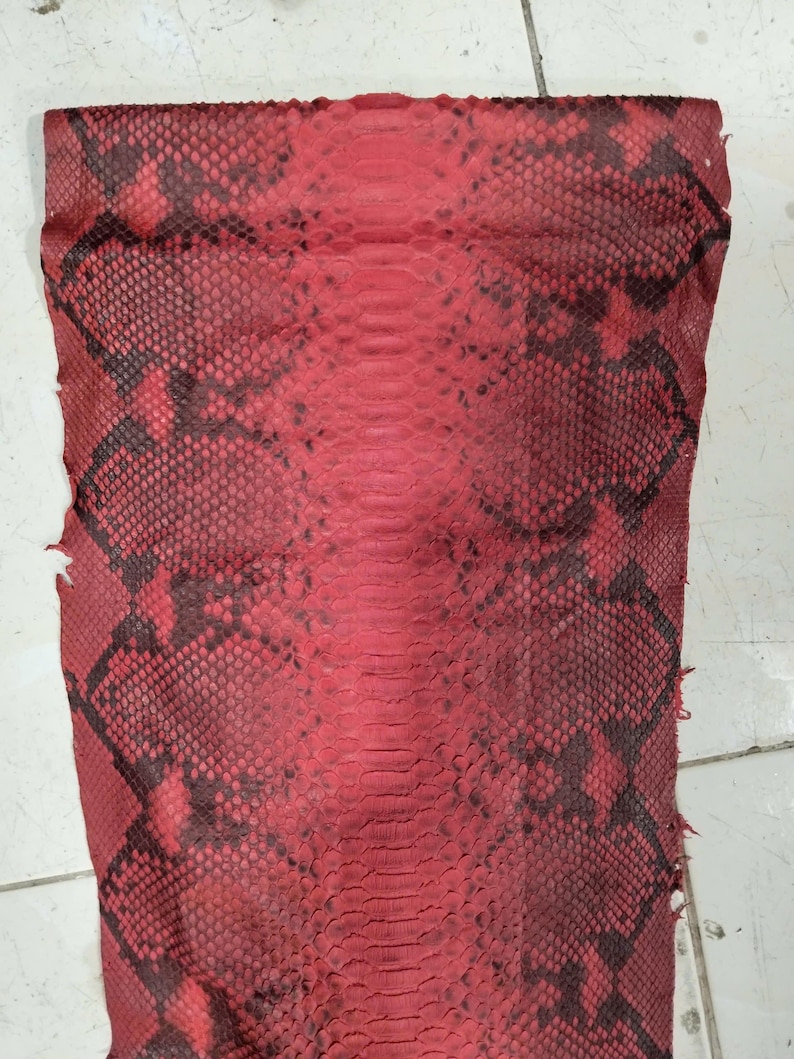 May include: A rectangular piece of red snakeskin leather featuring a distinct scale pattern with dark black markings. The hide displays a central row of larger, elongated scales flanked by smaller, textured scales across the entire surface area.
