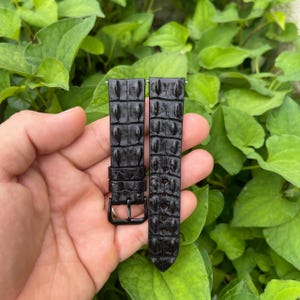 Puede incluir: Dos correas de reloj negras con textura de piel de cocodrilo. Las correas tienen forma rectangular con cierre de hebilla. El fondo es una vista borrosa de hojas verdes.