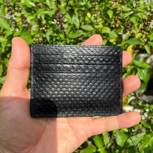 Puede incluir: Un tarjetero negro con un patr&oacute;n texturizado similar a la piel de serpiente. El tarjetero tiene m&uacute;ltiples ranuras para tarjetas y se sostiene en una mano contra un fondo de follaje verde.