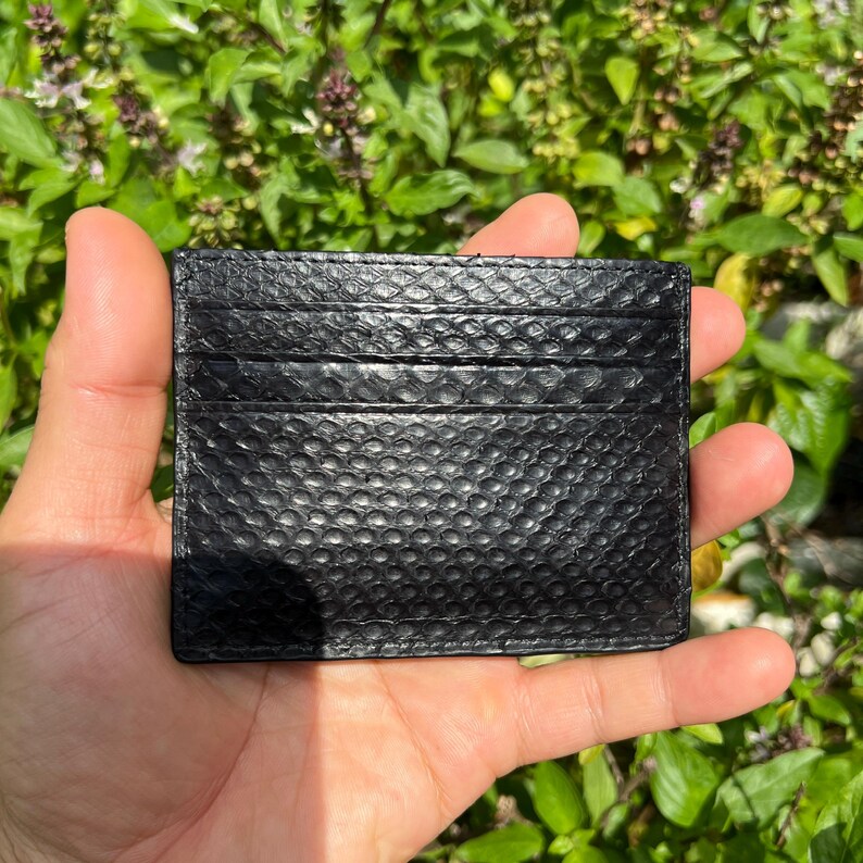 Puede incluir: Un tarjetero negro con un patr&oacute;n texturizado similar a la piel de serpiente. El tarjetero tiene m&uacute;ltiples ranuras para tarjetas y se sostiene en una mano contra un fondo de follaje verde.