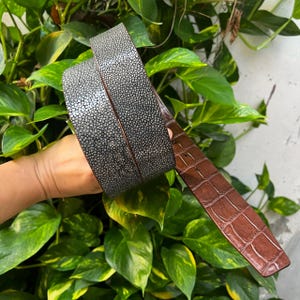 Puede incluir: Un cinturón con una textura de piel de raya gris y negra en la superficie exterior y un forro de cuero marrón liso con un acabado en relieve de cocodrilo en el interior, mostrando un contraste marcado entre ambos materiales.