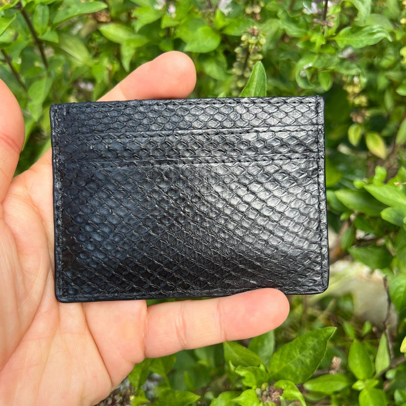 Puede incluir: Tarjetero negro con un patr&oacute;n texturizado de piel de serpiente. La cartera rectangular tiene m&uacute;ltiples ranuras para tarjetas y est&aacute; hecha de un material duradero. El tarjetero se sostiene frente a un fondo de hojas verdes.