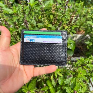 Puede incluir: Tarjetero de cuero negro texturizado con varias tarjetas de cr&eacute;dito visibles. El tarjetero se sostiene en una mano sobre un fondo de follaje verde. Las tarjetas tienen varios nombres y logotipos de empresas.