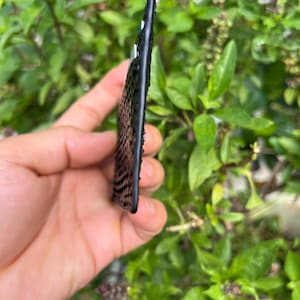 Puede incluir: Un objeto rectangular negro con una superficie texturizada, posiblemente una funda de tel&eacute;fono o un objeto decorativo. El objeto se sostiene en una mano sobre un fondo de follaje verde. La textura parece estar hecha de peque&ntilde;as lentejuelas oscuras.