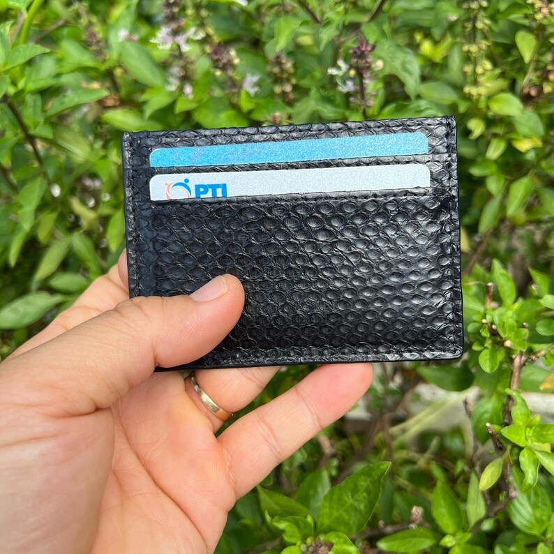 Puede incluir: Tarjetero de cuero negro texturizado con dos ranuras para tarjetas, que contiene dos tarjetas de cr&eacute;dito. El tarjetero se muestra frente a un fondo de hojas verdes.