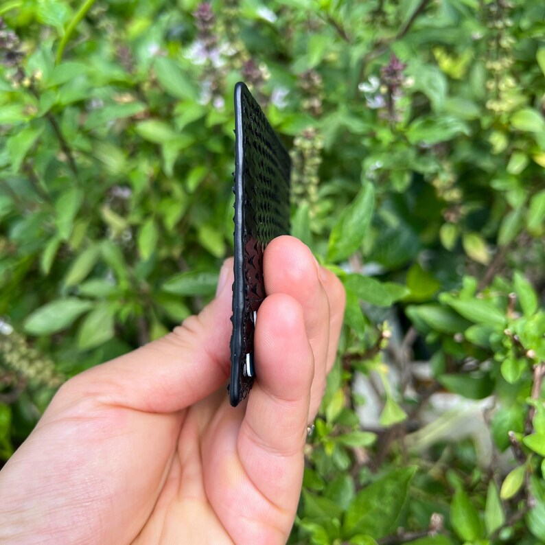 Puede incluir: Una tarjeta negra rectangular con una superficie texturizada. La tarjeta se sostiene frente a un fondo de follaje verde. Los bordes de la tarjeta son visibles, mostrando un patr&oacute;n ligeramente elevado.