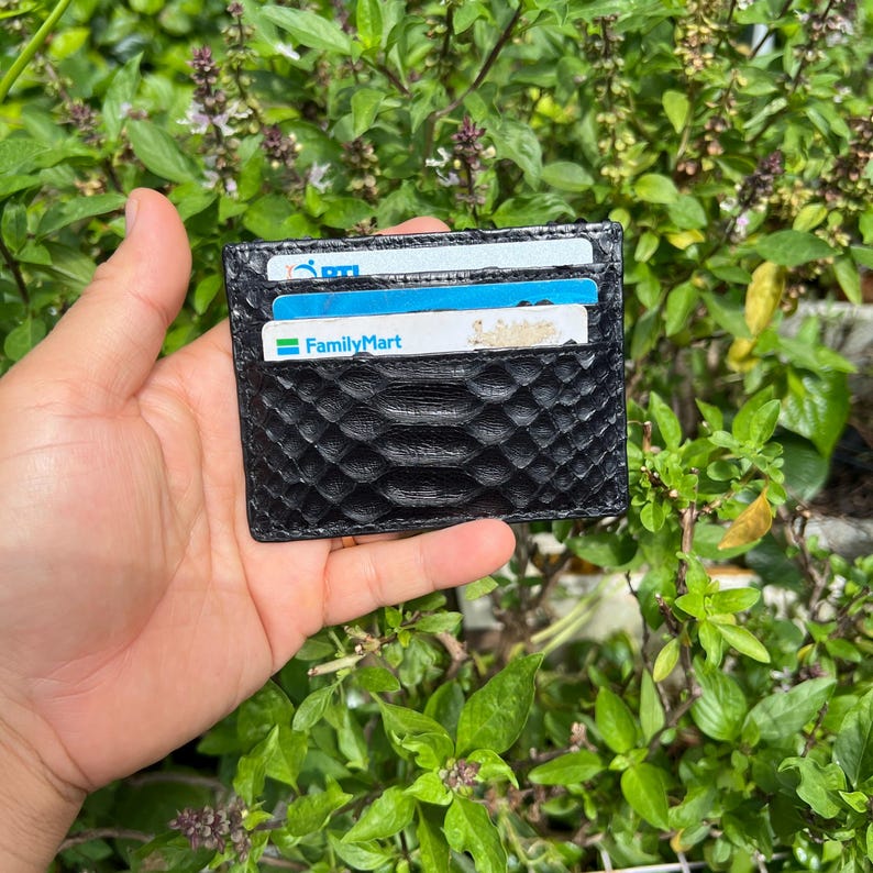 May include: A black cardholder with a textured, snake-like pattern. The wallet holds several cards, including one with the FamilyMart logo. The background features green foliage.