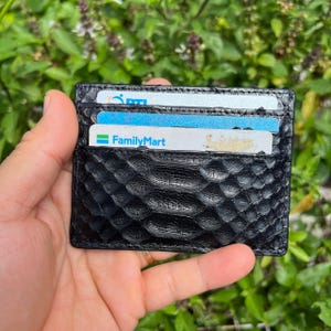 May include: Black leather card holder with a textured, snake-like pattern. Several cards are visible in the slots. The card holder is held in a hand against a background of green foliage.