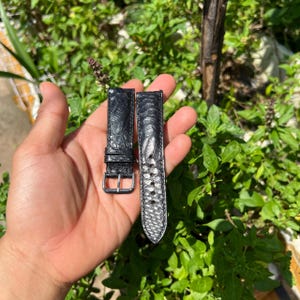 Może przedstawiać: Dwa czarne paski do zegarków z teksturowanym wzorem. Jeden pasek ma srebrną klamrę. Paski są prezentowane na tle zielonych liści.