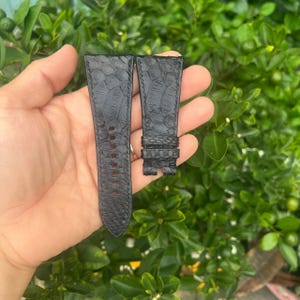 Può includere: Due cinturini neri per orologio con un motivo strutturato simile a quello di un rettile. Un cinturino ha più fori per la regolazione, l'altro ha una fibbia. I cinturini sono mostrati su uno sfondo di fogliame verde.