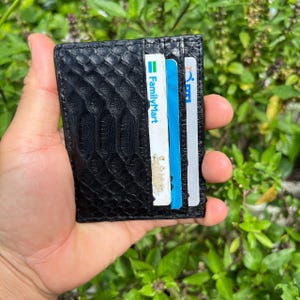 May include: A black cardholder with a textured snakeskin pattern. The wallet is open, revealing several cards with blue, white, and green accents. The FamilyMart logo is visible on one card. The wallet is held in a hand against a green leafy background.