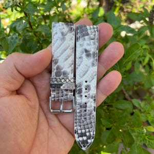 Puede incluir: Dos correas de reloj con un patrón de piel de serpiente en tonos blancos, grises y negros. Las correas tienen una hebilla plateada y se sostienen sobre un fondo de follaje verde.