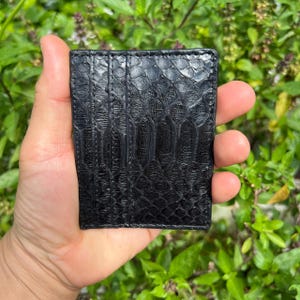 May include: A black, textured leather cardholder with a reptile-like pattern. The cardholder is rectangular and appears to be made of high-quality leather. The background is a blurred view of green foliage.