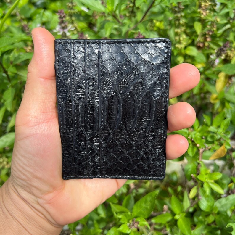 May include: A black, textured leather cardholder with a reptile-like pattern. The cardholder is rectangular and appears to be made of high-quality leather. The background is a blurred view of green foliage.
