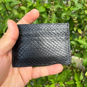 Puede incluir: Un tarjetero negro con un patr&oacute;n texturizado de piel de serpiente. El tarjetero tiene m&uacute;ltiples ranuras y se sostiene en la mano de una persona contra un fondo de follaje verde.