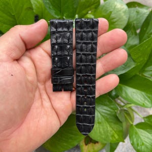 Puede incluir: Correa de reloj negra con un patrón texturizado similar al de un cocodrilo. La correa está hecha de cuero y tiene un cierre de hebilla. La correa del reloj se muestra sobre un fondo de hojas verdes.