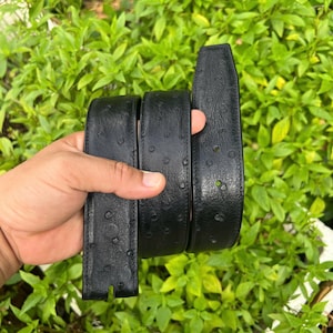 Puede incluir: Un cinturón de cuero negro con una superficie texturizada y una serie de pequeños agujeros para la hebilla. El cinturón se sostiene contra un fondo de follaje verde.