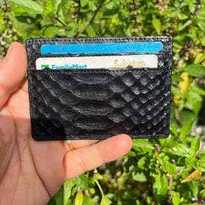 Könnte beinhalten: Schwarzer Kartenhalter mit einem strukturierten, schuppenartigen Muster. Das Portemonnaie hat zwei Kartenfächer, eines mit einer blauen Karte und das andere mit einer FamilyMart-Karte. Der Kartenhalter wird vor einem Hintergrund aus grünem Laub gehalten.