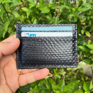Puede incluir: Tarjetero negro con un patr&oacute;n texturizado de piel de serpiente. La cartera es rectangular y contiene dos tarjetas, una con el texto "PTI". El tarjetero se sostiene en una mano sobre un fondo de hojas verdes.