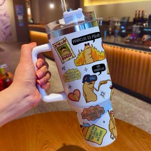 May include: White insulated tumbler with a handle, featuring cartoon graphics and text. The tumbler has a clear lid and a straw. The graphics include a character with the text "MARCUS IS FINE" and "VR MARCUS".