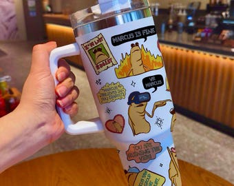 May include: White insulated tumbler with a handle, featuring cartoon graphics and text. The tumbler has a clear lid and a straw. The graphics include a character with the text "MARCUS IS FINE" and "VR MARCUS".