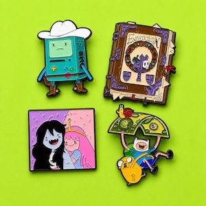 May include: Four enamel pins featuring Adventure Time characters and items: a teal BMO wearing a white cowboy hat, a brown Enchiridion book with text, a square pin showing Marceline and Princess Bubblegum, and Finn and Jake under a green leaf.