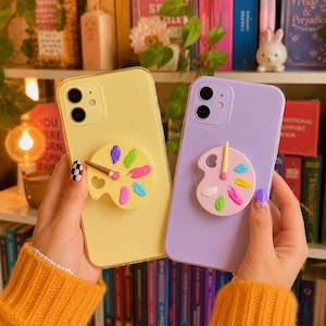May include: A pastel yellow smartphone case and a pastel lavender smartphone case, each featuring a circular paint palette grip on the back. The yellow case has a palette with a brown-handled brush, while the lavender case has a pink-handled brush.