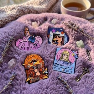 Puede incluir: Cuatro pines de esmalte con diseños ilustrados: una figura con vestido rosa y guitarra azul, un retrato con el texto Midnights y estrellas, una figura de cabello rubio con el texto Lover y una escena desértica con el texto The Life of a Showgirl.