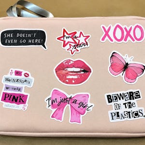 May include: A light pink rectangular laptop sleeve decorated with various stickers including a pink butterfly, red lips, a pink bow, and text phrases such as "She doesn't even go here," "XOXO," "On Wednesdays we wear pink," "I'm just a girl," and "Beware of the plastics."